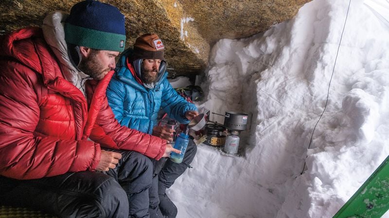 Two people bundled up in a snow cave with a camp stove and cookwear. One person pours water from a sierra cup into the other person’s water bottle.&nbsp;